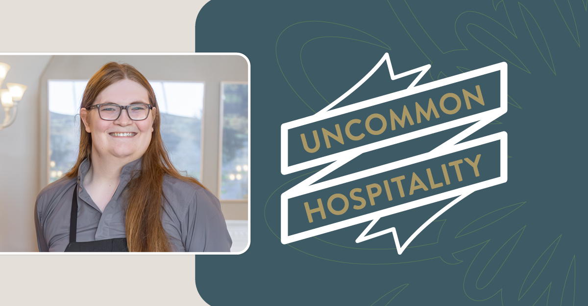 Portrait of Elle, who has brown hair and glasses, smiling. A banner motif to the right says "Uncommon Hospitality"