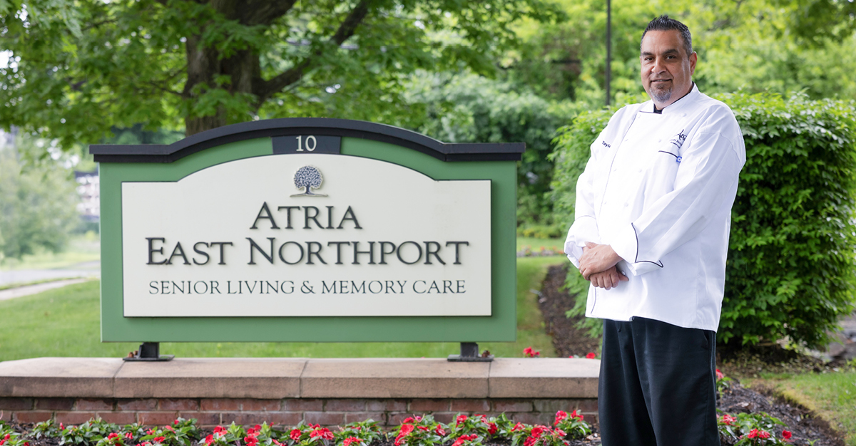 Chef Peter Taylor stands outdoors beside the Atria East Northport Senior Living & Memory Care sign.